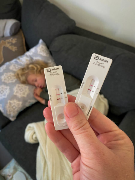 Two COVID positive tests shown in front of a sleeping, sick toddler