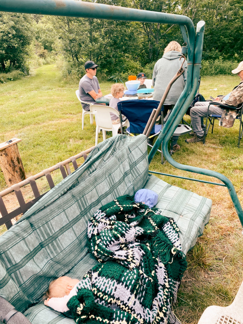 a baby sleeps on an ourdoor swing as a family prepares for dinner in the background while camping in the summer on cape breton island